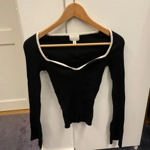 Svart ribbad topp med vit kant H&M - Svart långärmad ribbad topp från H&M med vit kant runt den breda, något båtformade urringningen. Toppens material är stretchigt och sitter tight. Perfekt för dig som gillar minimalistisk stil med en twist.
