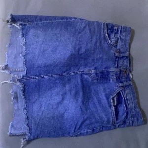Blå jeanskjol med fransad kant från 157 - Säljer en blå jeanskjol från 157 med fransad och rå kant nedtill. Kjolen har klassiska fickor fram och bak, samt knappar i midjan. Perfekt för dig som gillar en chill och avslappnad stil med lite edge.