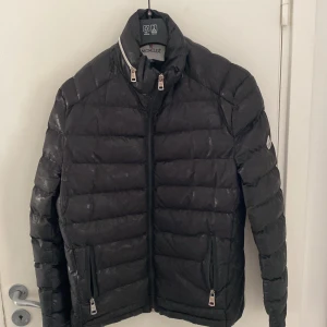 Svart Moncler dunjacka herr - Snygg svart dunjacka från Moncler med quiltad design och hög krage. Jackan har dragkedja framtill, två fickor med dragkedja och diskret Moncler-logga på ärmen. Perfekt för kalla dagar och ger en clean, stilren look.