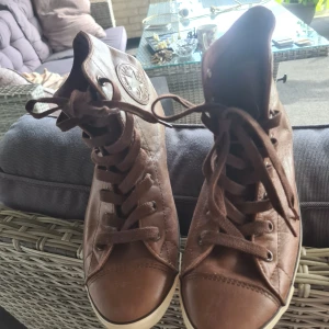 Bruna Converse All Star high-tops stl 38 - Säljer ett par snygga bruna Converse All Star high-tops i skinn med klassisk rund tå och platt sula. Skorna har snörning och det ikoniska Converse-märket är präglat på sidan. Perfekt för dig som vill ha en cool och tidlös look.