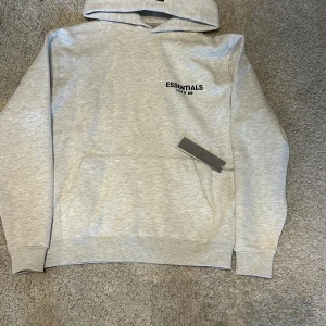 Grå Essentials Fear of God hoodie - Säljer en ljusgrå hoodie från Essentials Fear of God med svart logga på bröstet och ryggen. Tröjan har huva, känguruficka och ribbade muddar. Materialet är mjuk bomull och polyester, perfekt för chill dagar. Stilren och enkel design som funkar till streetwear.
