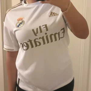 Real Madrid vit fotbollströja Adidas - Snygg vit Real Madrid fotbollströja från Adidas med guldiga detaljer och klubbmärke på bröstet. Tröjan har korta ärmar, guldiga ränder på axlarna och Emirates-logga framtill. Tillverkad i lätt och ventilerande material, perfekt för match eller träning. Barn storlek L 164 passar perfekt för en Xs.