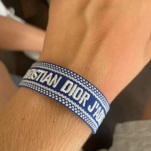 Blått Dior armband med tofsar - Snyggt blått armband från Christian Dior med vit broderad text 'J'ADIOR'. Armbandet har ett coolt mönster och knytning med två tofsar i blått och vitt. Perfekt accessoar för att lyfta din outfit med lite lyxig vibe.