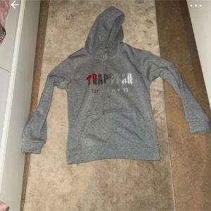 Grå hoodie Trapstar med tryck - Säljer en grå hoodie från Trapstar med tryck i svart, vitt och rött framtill. Tröjan har huva med snörning och en stor magficka. Perfekt för dig som gillar streetwear och vill ha en chill look.