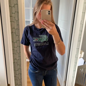 Mörkblå Lacoste t-shirt - Säljer en mörkblå t-shirt från Lacoste med den klassiska loggan på bröstet. T-shirten är i storlek 176 men jag är 162 och den passar.