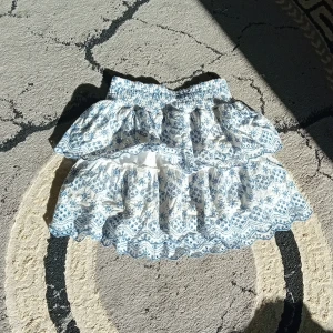 Vit och blå volangkjol med mönster - Supersöt kort kjol med volanger och blått blommigt mönster på vit botten. Kjolen har bred resår i midjan och två lager med volanger som ger en fluffig och lekfull look. Perfekt för dig som gillar romantisk och feminin stil.