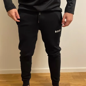 Svarta mjukisbyxor från BOSS, strl S - Svarta mjukisbyxor från BOSS i storlek S med vit logga på vänster lår. Byxorna har snörning i midjan och ribbade muddar vid bensluten. Skön passform som sitter snyggt och är perfekt för chill eller träning. Materialet är mjukt och stretchigt för maximal komfort.