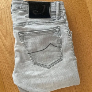 Jacob Cohen Jeans - Snygga ljusgrå jeans från Jacob Cohen med klassisk femficksdesign och svart patch med logga bak. Jeansen har smal passform och är tillverkade i mjukt denimtyg som sitter riktigt bra. Perfekta för dig som gillar stilrena och moderna jeans. De är ett litet håll i skrevet. Priset är inte skrivet i sten!! 