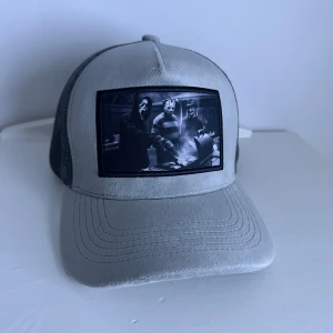 Grå grisch keps  - Snygg grå keps med svart mesh på baksidan och justerbar snapback. Framtill finns ett svartvitt patch med motiv från klassiska skräckfilmer, bland annat Ghostface och Jason. Perfekt för dig som gillar film eller vill sticka ut med en unik accessoar.