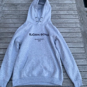 Grå hoodie från Björn Borg - Snygg grå hoodie från Björn Borg med svart tryck på bröstet och texten 'Stockholm 1956'. Tröjan har huva med snörning och en stor magficka framtill. Perfekt för chill dagar och enkel att matcha med jeans eller joggers.