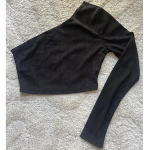 Svart oneshoulder topp  - Snygg svart croppad topp med ribbad struktur och oneshoulder-design. Toppen har en lång ärm på ena sidan och är axelbandslös på andra, vilket ger en cool asymmetrisk look. Perfekt att styla med höga jeans eller kjol för en trendig vibe.