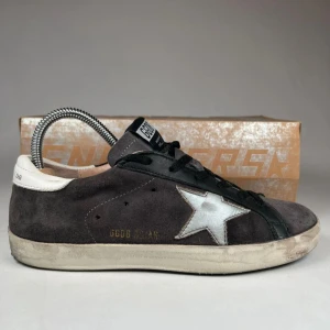 GGDB sneakers med silverstjärna - Snygga GGDB sneakers som passar 40 i mörkgrå mocka med svart snörning och vit häl. På sidan finns en stor silverfärgad stjärna som ger skorna en unik vibe. Sulan är vit och har en vintage-look. Perfekta för dig som gillar streetstyle och italiensk design.