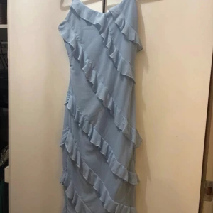 Ljusblå långklänning med volanger - Säljer en ljusblå långklänning med offshoulder-design och flera diagonala volanger som ger en cool och romantisk vibe. Klänningen har en flowy passform och är perfekt för sommarens alla tillfällen. Materialet känns lätt och luftigt.