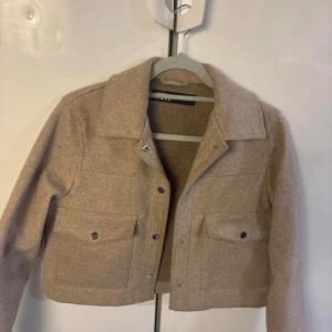 Beige overshirt jacka från Zara - Snygg beige overshirt jacka från Zara i storlek XS. Jackan har en croppad passform, stora fickor med knappar framtill och klassisk krage. Materialet är mjukt och känns som ull, perfekt för lager på lager. Enkel och stilren design som funkar till många outfits.