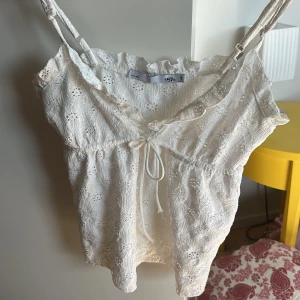 Vit broderad linnetopp med knyt - Supersöt vit linnetopp från 157 med broderade blommor, smala axelband och v-ringning. Toppen har volangkant upptill och knytband framtill för extra detalj. Perfekt till jeans eller kjol för en fräsch sommarlook!