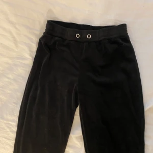 Svarta mjukisbyxor i sammet - Svarta mjukisbyxor i mjuk sammet med bred resår i midjan och metallöljetter framtill. Perfekta för chill dagar eller när du vill vara extra bekväm. Klassisk rak passform och enkel stil som funkar till det mesta.