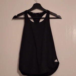 Svart linnetopp från Adidas - Svart linnetopp från Adidas med klassisk racerback och logga framtill vid nederkanten. Toppen är ärmlös och har en sportig look, perfekt för träning. Tillverkad i mjukt och lätt material som andas.