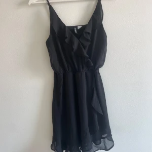 Svart kort klänning  - Säljer en svart kort klänning från H&M Divided i storlek 34. Klänningen har justerbara smala axelband, v-ringning med volangdetalj framtill och ett lätt, flödande tyg som ger en luftig känsla. Perfekt för dig som gillar enkel men snygg stil. Aldrig använd