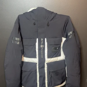 Sail Racing glacier dunjacka - Säljer en fet svartvit dunjacka från Sail Racing med stora fickor, huva och coola loggor på ärmarna. Jackan har en schysst blockdesign och dragkedja framtill. Perfekt för kalla dagar när du vill se både sportig och stilren ut.