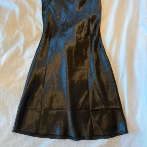 Svart satin klänning med öppen rygg - Snygg svart klänning i glansigt satinmaterial med smala axelband och en cool öppen rygg med snörningsdetaljer. Klänningen har en figurnära passform och är kort i modellen, perfekt för dig som gillar trendiga och stilrena plagg.