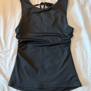 Svart topp med öppen rygg - Säljer en svart topp med breda axelband och snygg öppen rygg med knytning. Modellen är enkel och stilren. Materialet är stretchigt och bekvämt, vilket gör den lätt att röra sig i.