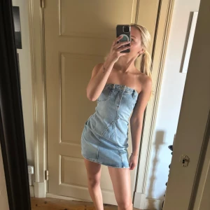 Ljusblå jeansklänning axelbandslös - Säljer en snygg axelbandslös jeansklänning i ljusblå denim. Klänningen är kort och har en tight passform med coola sömdetaljer och knapp framtill. Perfekt för dig som gillar en trendig och avslappnad stil.