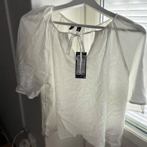 Vit blus med knyt från Vero Moda - Fräsch vit blus från Vero Moda i storlek S. Blusen har korta puffärmar och en rundad halsringning med snygg knytning framtill. Materialet är lätt och luftigt, perfekt för ljusa dagar. Enkel och stilren design som funkar till många looks.