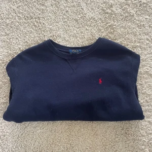 Mörkblå sweatshirt från Polo Ralph Lauren - Snygg mörkblå sweatshirt från Polo Ralph Lauren med klassisk rund hals och långärmad passform. Tröjan har det ikoniska röda Polo-logot broderat på bröstet och är gjord i mjuk bomull. Perfekt för chill dagar och enkel att matcha med jeans eller joggers.