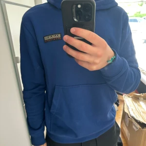 Blå hoodie från Napapijri - Säljer en blå hoodie från Napapijri med klassisk logga på bröstet. Tröjan har huva med snörning, stor magficka och långa ärmar. Perfekt för chill dagar och enkel att matcha med jeans eller joggers. Skön och mjuk bomullskvalitet. Jag är 181cm och 75kg