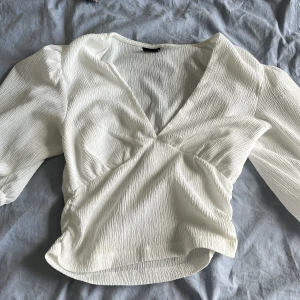 Vit omlottblus från Gina Tricot - Supersnygg vit omlottblus från Gina Tricot i storlek S. Blusen har v-ringning, långa puffärmar och är gjord i ett lätt, krinklat tyg som ger en cool struktur. Perfekt att styla med jeans eller kjol för en fräsch look.