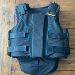 Svart säkerhetsväst från Airowear - Säljer en svart säkerhetsväst från Airowear, modell Junior Outlyne. Västen har blå detaljer och justerbara kardborreband i midjan och axlarna. Materialet är slitstark syntet med mesh på insidan för extra komfort. Följsam model som blir rörlig och formad av ryttaren när den används ett tag. Perfekt för ridning och ger bra skydd. Använd fåtal gånger, inga avramlingar. Säljs då den är för liten. Köptes för ca. 1700kr. Pris går att förhandla.