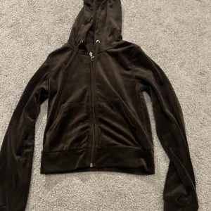 Mörkbrun velour hoodie från Gina Tricot - Säljer en mörkbrun hoodie från Gina Tricot i mjuk velour med dragkedja och huva. Tröjan har snörning vid huvan, två fickor fram och en snygg ringdetalj på dragkedjan. Perfekt för chill dagar och sköna outfits.