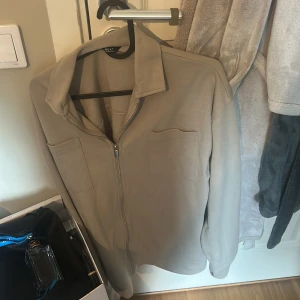 Beige overshirt från Next med dragkedja - Snygg beige overshirt från Next med dragkedja framtill och två bröstfickor. Enkel och clean design med klassisk krage, perfekt att slänga över en t-shirt. Mjuk känsla och passar till många olika outfits. Oanvänd