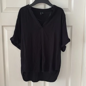 Svart v-ringad blus från Vero Moda  - Svart blus från Vero Moda i storlek L med v-ringad hals och korta ärmar. Blusen har en avslappnad passform, uppvikta ärmslut och något längre bakstycke. Perfekt för dig som gillar enkel och stilren design. 