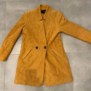 Senapsgul kappa från Stradivarius - Snygg senapsgul kappa från Stradivarius i klassisk modell med två knappar framtill och fickor på sidorna. Jackan har lång ärm och är tillverkad i ett mjukt tyg med struktur. Perfekt för dig som vill sticka ut med färg i höst och vinter.