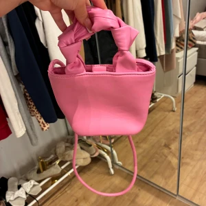 Rosa axelväska med knutdetalj - Söt liten axelväska i bubbelgumsrosa syntetmaterial. Väskan har ett handtag med knutdetaljer och en lång axelrem. Perfekt storlek för mobilen och det viktigaste. Stängs med magnetknapp och har ett fack inuti. Superfin accessoar för att liva upp din outfit.