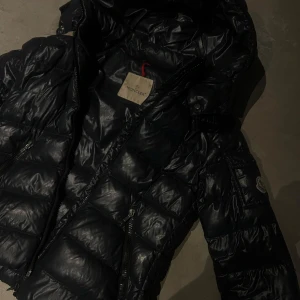 Svart/blå  Moncler pufferjacka med huva - Nu säljer jag min svart/blå moncler jacka då den blivit förliten. Den har använts mycket och har några defekter. Lappen har gått av och den har ett hål längst ner på vänstra armen, men det går att fixa hos skräddare. Den är köpt på nk. Den är i barnstorlek men står ingen storlek man jag skulle säga 12-13 jag själv är 162 lång så om man vill se hur den sitter på mig kan man skriva privat då jag inte fick plats med fler bilder.samma gäller mått. Pris kan sänkas!!! Den är i äkta dun