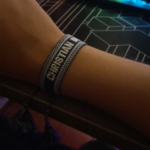 Dior armband  - Snyggt blått armband från Christian Dior med texten 'J'Adior' broderad i vitt. Armbandet har en bred, vävd design med vita detaljer och justerbara snören med tofsar. Perfekt för dig som vill ha en cool designeraccessoar.