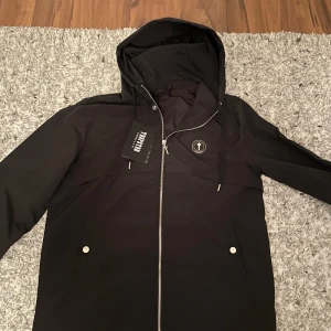 Svart jacka från Trapstar - Säljer en svart jacka från Trapstar med huva och dragkedja. Jackan har en cool logga på ryggen och praktiska fickor med knappar framtill. Perfekt för en stilren look.