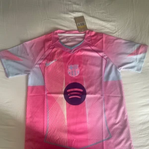 FC Barcelona rosa matchtröja Nike - Snygg FC Barcelona fotbollströja från Nike i rosa och ljusblått med vita linjer och klubbmärke på bröstet. Tröjan har korta ärmar, rund hals och är gjord i lätt, ventilerande material. Spotify-logga på magen och modern design som sticker ut på planen.