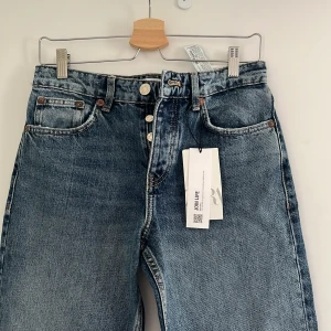 Blå raka jeans från Zara, strl 36 - Snygga blå jeans från Zara i rak modell med klassisk femficksdesign och knäppning med knappar. Jeansen har en cool tvättad look och är tillverkade i bomull. Perfekta för dig som gillar en avslappnad och tidlös stil. Jeansen är aldrig använda och prislappen fortfarande kvar 