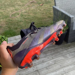 Adidas Predator fotbollsskor grå/röd - Säljer ett par Adidas Predator fotbollsskor i grått och svart med röda detaljer och sula. Skorna har snörning och är designade för gräsplan, med mönstrad ovandel och klassiska Predator-loggor. Perfekta för dig som vill sticka ut på planen.