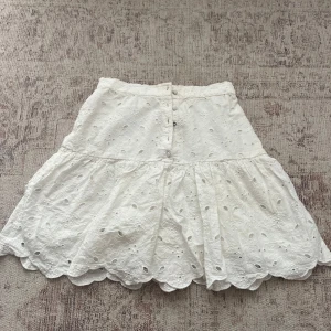 Vit broderad kjol från Holly & Whyte - Supersöt vit kortkjol från Holly & Whyte med broderade blommor och spetsdetaljer. Kjolen har knappar framtill, volangkant och är gjord i mjuk bomull. Perfekt för sommarens alla dagar, matcha med en enkel topp eller hoodie för en chill look. En av knapparna är borta och därav det billiga priset, fortfarande en väldigt fin kjol, pris kan diskuteras vid snabb affär💞