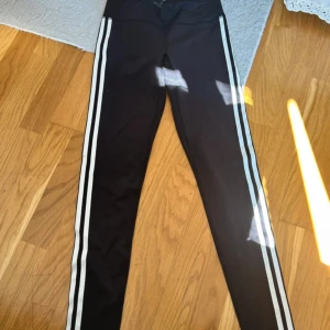 Svarta Adidas leggings med vita ränder - Svarta leggings från Adidas med klassiska vita ränder längs sidorna. Tight passform och hög midja, perfekta för träning eller chill. Tillverkade i stretchigt syntetmaterial som sitter skönt och följer kroppen. Adidas-logga framtill i midjan.