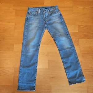 Levis 501 ljusblå jeans - Säljer ett par Levis 501 jeans i ljusblå tvätt. Klassisk femficksmodell med raka ben och normal passform. Jeansen har snygga slitningar och är tillverkade i mjukt bomullsjeansmaterial. Perfekta för en avslappnad och stilren look. Jeansen är endast använda en gång, säljes då ja inte riktigt älskade hur de satt på mig. 31 i Midjan och 32 i längd