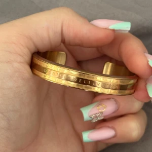 Guldigt armband från Daniel Wellington - Snyggt och stilrent armband i guldton från Daniel Wellington. Armbandet är öppet och har en graverad logga längs kanten. Designen är enkel men ändå lyxig med en slät, blank yta och diskreta cirkeldetaljer. Perfekt för dig som gillar klassiska accessoarer med en modern touch.