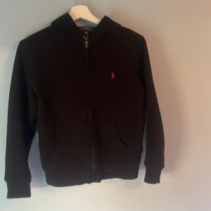 Svart hoodie från Polo Ralph Lauren - Svart hoodie från Polo Ralph Lauren med röd broderad logga på bröstet. Tröjan har huva, dragkedja framtill och två fickor. Tillverkad i mjuk bomull och har långa ärmar. Perfekt för chill dagar och streetwear-stil. 10-12 och inga defekter annars än ett litet hål i luvan som knappt syns