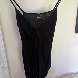 Svart topp från Gina Tricot - Svart topp med smala axelband och snygg knytdetalj framtill. Klänningen har en v-ringad urringning och är figurnära med en stilren look. Perfekt för dig som vill ha en enkel men chic outfit.