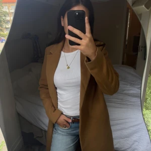 Klassisk brun kappa med krage - Säljer en snygg, lång brun kappa med klassisk krage och knäppning framtill. Jackan har en rak passform och är perfekt för dig som gillar stilrena och tidlösa plagg. Passar till jeans eller dressat och är superenkel att matcha.