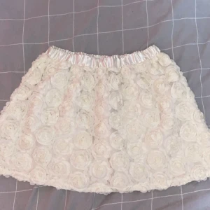 Vit blommig kortkjol från Lindex - Supersöt vit kortkjol från Lindex med 3D-blommor över hela tyget och glansig resår i midjan. Kjolen har en fluffig look och passar perfekt när du vill sticka ut med en unik och romantisk stil. Perfekt för dig som gillar detaljer och blommigt.